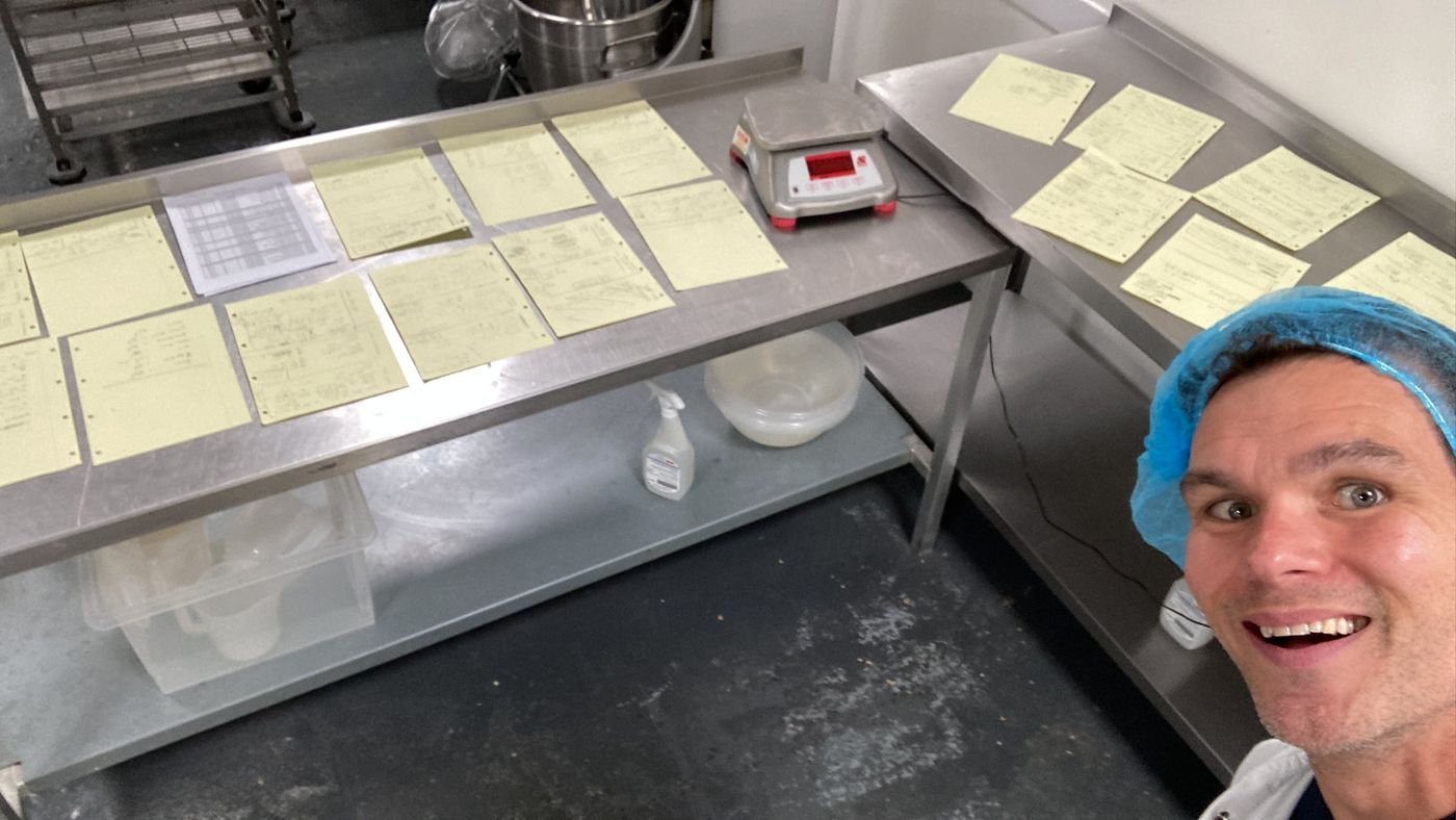 Ro in a hairnet grinning beside recipe sheets and scales on a factory bench